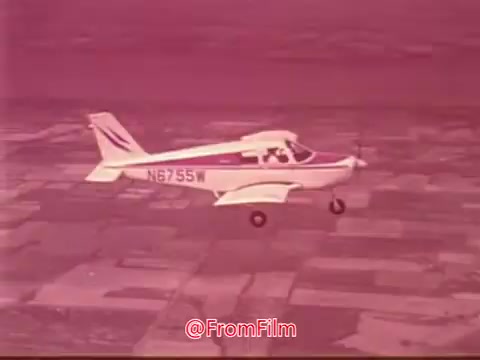 The still depicts a small aircraft, specifically a Cessna, in flight. The plane is shown from a slightly elevated angle as it approaches a patchwork landscape below. The image has a vintage look typical of 16mm film, possibly featuring a reddish hue. This scene is likely part of a Public Service Announcement (PSA) where Jimmy Stewart, known for his advocacy for the Civil Air Patrol, promotes aviation and community service.