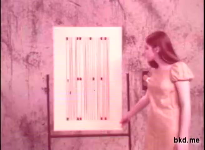 The still depicts a woman in a 1970s-style dress standing beside a poster on an easel. The poster features a series of vertical lines and dots, likely used for a visual demonstration. The color tones are muted, with a pinkish hue prevalent in the image, characteristic of 16mm film prints from that era. The background appears textured and simple, emphasizing the educational nature of the scene.