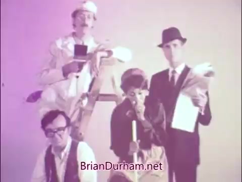 The still from the PSA 'A Matter of Life and Breath' features a group of individuals in a stylized, playful presentation. At the forefront, a woman holds a broom and appears contemplative, representing the 'Sad Commuter' theme. Behind her, three men, including James Noble, are dressed in various outfits, adding to the humorous yet serious tone of the public service announcement. The image is visually striking with a vintage color palette typical of the 60s and 70s, conveying a message about respiratory health in a memorable, engaging way.