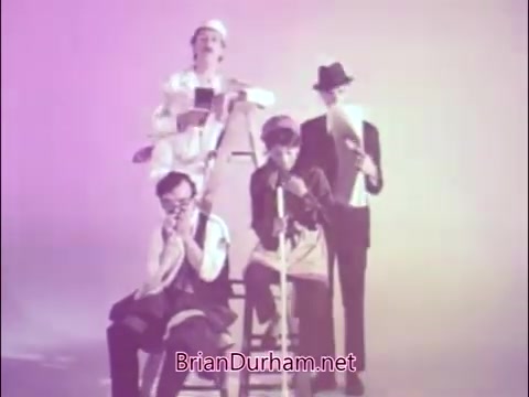 The still depicts a group of four men in various comedic poses, highlighting a humorous yet poignant theme related to commuting. One man stands on a ladder, possibly depicting an elevated perspective, while the others are seated, immersed in their daily routines. The color palette seems soft and muted, creating an almost dreamlike quality. The context is a public service announcement from the National Tuberculosis and Respiratory Disease Association, aiming to raise awareness about respiratory health. This PSA features James Noble, known for his role as 'Sad Commuter,' capturing the struggles of daily life while subtly linking it to respiratory health issues.