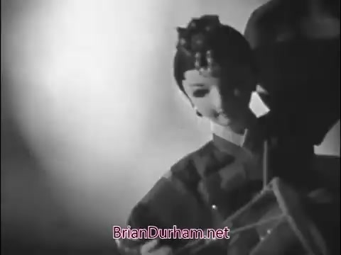 The still features a vintage 16mm black-and-white scene from a 1960s Girl Scout commercial. It shows a stylized, possibly animated, character resembling a girl in traditional attire, likely promoting themes of community or friendship. The artistic lighting creates a dramatic effect, emphasizing the character's expression and attire. The overall mood reflects the era's charm and messaging of unity among girls in scouting.