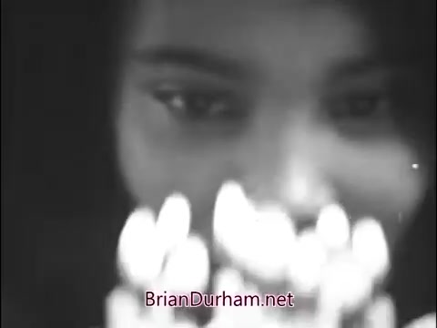 The still from the 1960s Girl Scout commercial features a young girl with a focused expression, gazing intently at a cluster of lighted candles. The soft lighting emphasizes her features and the glow of the candles, evoking a sense of warmth and community spirit associated with the Girl Scouts. The overall mood appears reflective and hopeful, capturing the essence of childhood and collective experiences.