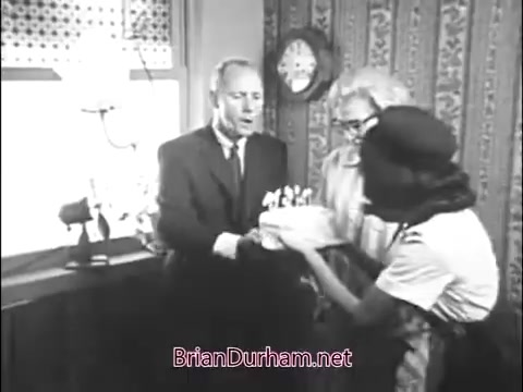 The still shows two men and a Girl Scout in a vintage setting, likely from the 1960s. The Girl Scout, dressed in a uniform, is presenting a cake to one of the men, who appears surprised or pleased. The background features a patterned wallpaper and a decorative clock, suggesting a warm, community-oriented atmosphere typical of the era. The scene encapsulates themes of camaraderie and celebration within the community.