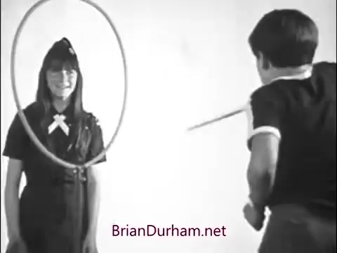 The still features a girl dressed in a Girl Scout uniform with a big smile, standing behind a large hoop. A boy is positioned in front of her, seemingly about to jump through the hoop. The scene reflects a playful, community-oriented atmosphere typical of 1960s children's commercials.