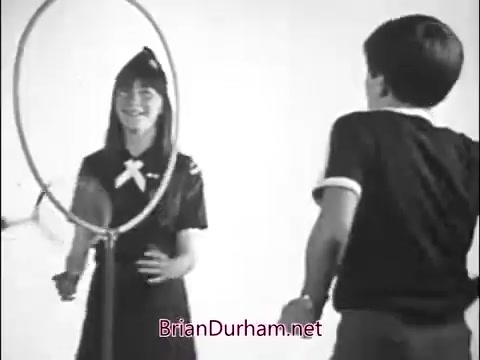 The still from the 1960s Girl Scout commercial depicts a young girl wearing a Girl Scout uniform and a boy engaging in a playful interaction involving a large hoop. The girl has a joyful expression, indicating a fun, friendly atmosphere. The backdrop appears to be simple, enhancing the focus on their activity. The scene captures the spirit of community and camaraderie typical of Girl Scout promotions from that era.