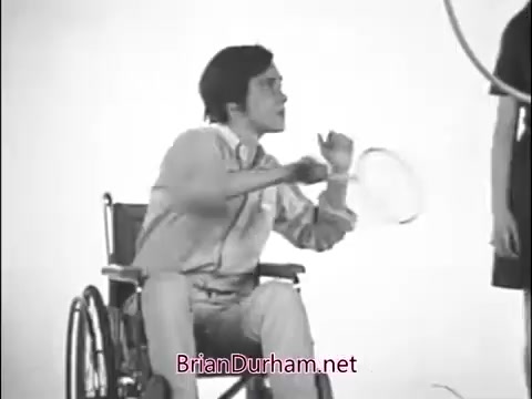 The still shows a young man in a wheelchair, engaged in a lively interaction with a girl. He holds a racquet, possibly indicating a game or sport that promotes inclusivity and community. The backdrop is simple, emphasizing the focus on the characters. This scene reflects the themes of empowerment and camaraderie often highlighted in Girl Scout commercials from the 1960s.