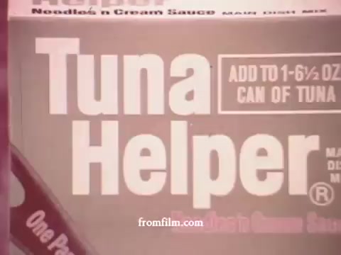 The still features a close-up of a Tuna Helper box from the 1970s. The packaging prominently displays the 'Tuna Helper' name in bold, white letters against a pink background. Instructions indicate the product is to be combined with a specified amount of canned tuna. The design reflects the era's aesthetic, with simple graphics typical of processed food advertisements from that time.