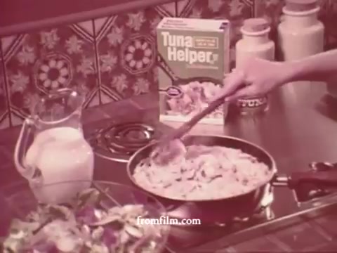 The still features a vintage kitchen scene typical of the 1970s. A hand stirs a pot on the stove, containing a creamy mixture, possibly the Tuna Helper dish being prepared. In the background, a box of Tuna Helper is prominently displayed, accompanied by a glass jug, likely containing milk, and a bowl of fresh salad. The decor reflects the era's style, with patterned tiles adding to the nostalgic feel.