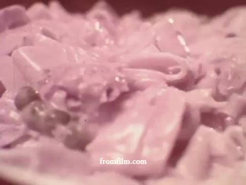 The still captures a close-up of a creamy pasta dish featuring a mixture of tuna, noodles, and peas, all presented in a pinkish sauce. The texture appears rich and hearty, characteristic of the era's comforting meal preparations. The overall aesthetic reflects a vintage 1970s style typical of food advertisements from that time.