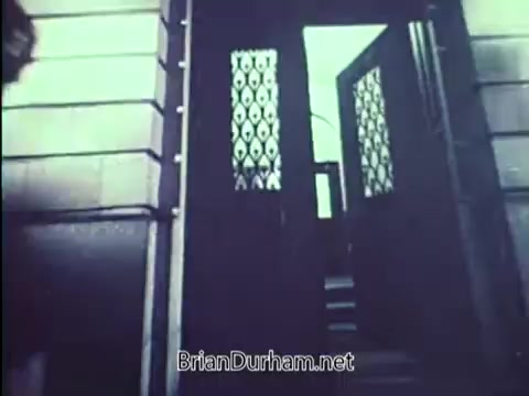 The still from the 1967 PSA 'Lock Your Car' by the United States Department of Justice features a doorway, possibly to a building. The door is partially open, revealing an interior staircase. The door has a decorative design, indicating a formal or institutional setting. The image likely aims to convey a message about security and the importance of safeguarding one's belongings, as is central to the PSA's theme.