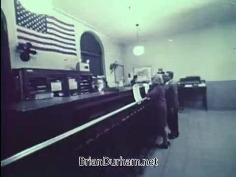 The still from the 1967 PSA by the United States Department of Justice features a scene inside a government building. In the foreground, two individuals approach a counter, likely engaged in a conversation or reporting an incident. Behind them, there is an American flag prominently displayed on the wall, adding a patriotic context to the setting. The room appears functional and orderly, reflecting the seriousness of the message about car safety and crime prevention.