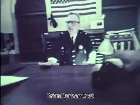 The still from the 1967 PSA 'Lock Your Car' features a middle-aged man, likely a police officer or official, seated at a desk. He wears a uniform adorned with badges, glasses, and gestures as he speaks. Behind him is an American flag, adding to the authoritative context of the message aimed at promoting vehicle security. The setting appears formal, suggesting a serious tone.