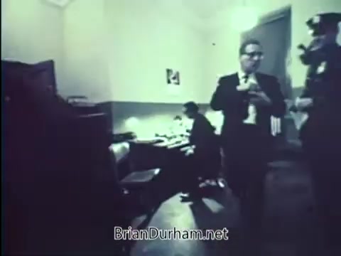 The still from the 1967 United States Department of Justice PSA 'Lock Your Car' depicts a dimly lit office or room setting. In the foreground, a well-dressed man adjusts his tie while walking, and behind him, another individual is seated at a desk, possibly engaged in paperwork. A uniformed officer stands nearby, suggesting a formal or serious context, likely related to crime prevention or law enforcement. The overall ambiance conveys a sense of urgency regarding vehicle security.