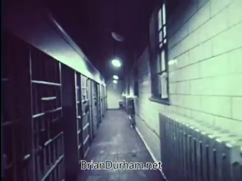 The still from 'United States Department of Justice PSA - Lock Your Car - 1967' features a dimly lit corridor, reminiscent of a prison setting, with jail cells on one side. The walls are bare and brick-like, illuminated by overhead lights that cast a stark glow, emphasizing the starkness and potential danger. This visual suggests themes of incarceration and consequences, reinforcing the importance of vehicle security to avoid theft.