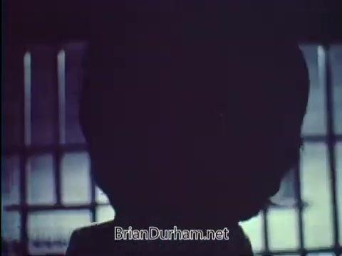 The still depicts a shadowy figure, likely a person, against a dimly lit backdrop with barred windows, suggestive of confinement or a secure environment. The visual tone is somber, aligning with the public service announcement's theme promoting car safety, specifically emphasizing the importance of locking vehicles to prevent theft. The ambiance conveys a serious message about the consequences of neglecting this simple precaution.