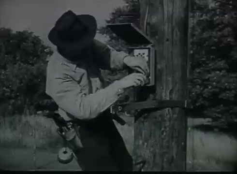 The still from 'Bell System Film - Drop Wire Runs Part 1 (b)' depicts a technician in a hat working on a utility pole. He is focused on a box attached to the pole, likely connecting or maintaining telephone wires. The surroundings appear rural, with greenery visible in the background. This scene is part of a film illustrating the process of telephone wire setup, highlighting the technical aspects of connecting the service from poles to customers.