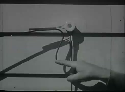 The still from 'Bell System Film - Drop Wire Runs Part 1 (b)' shows a hand pointing at a telephone drop wire installation on a building's exterior. The wire is attached to a fixture, likely designed to guide the wire from the pole to the customer, illustrating part of the telephone installation process. The simplicity of the setup reflects the practical techniques used in the late 1940s to early 1950s telecommunications.