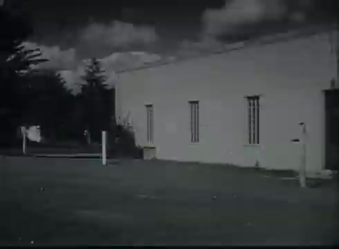 The still from 'Bell System Film - Drop Wire Runs Part 1 (b)' depicts a plain building with small windows, surrounded by a grassy area. The structure appears to be part of a Central Office facility, characteristic of late 1940s to early 1950s architecture. A clear sky with scattered clouds provides the backdrop, suggesting a calm day. The setting emphasizes the utilitarian nature of telephone infrastructure as it transitions from the pole to customer connections.
