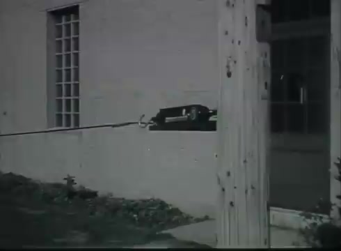 The still shows a section of telephone wire being installed from a pole to a building. The wire is secured to a device, possibly a puller or tool used in the installation process. The background features a simple wall with a window and a wooden pole, indicating an outdoor setting typical of utility installations. This visual underscores the practical steps involved in connecting telephone services.