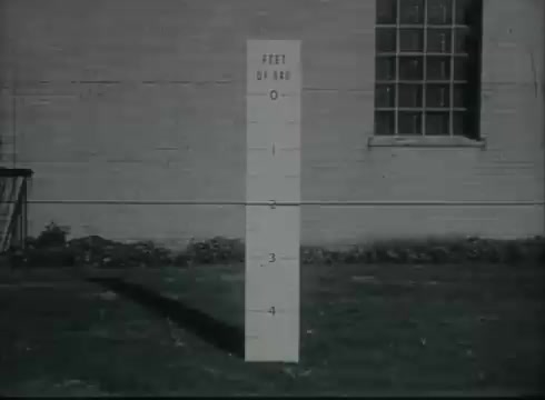 The still features a vertical measurement post marked with increments indicating feet, labeled 'FEET OF WIRE.' The post, set against a wall of a building with a window, serves as a visual reference for demonstrating the distance of telephone wire runs in the Bell System's instructional film from the late 1940s to early 1950s. The focus is likely on teaching about drop wire installations from telephone poles to customers.