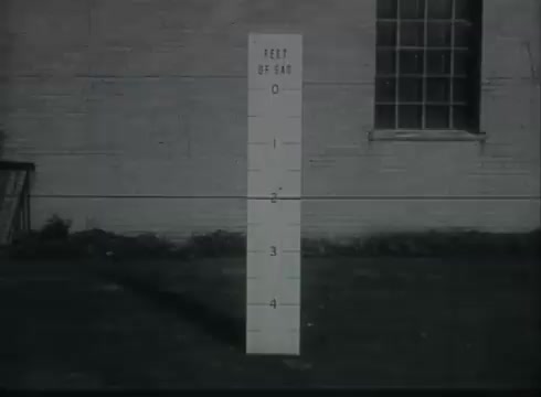 The still from 'Bell System Film - Drop Wire Runs Part 1 (b)' depicts a vertical, white measuring post marked in feet, showing increments from 0 to 4 feet. The post is situated in front of a wall with a window, likely in an outdoor setting. This visual aids viewers in understanding height measurements relevant to telephone wire installation from utility poles to customer premises, conveying technical information in a straightforward manner.