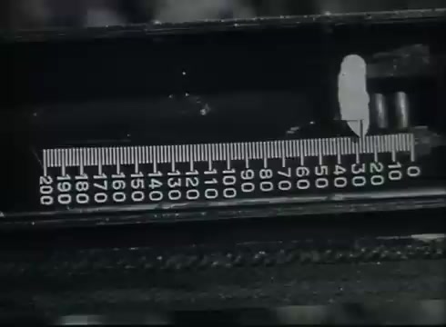 The still from 'Bell System Film - Drop Wire Runs Part 1 (b)' shows a close-up of a measuring device, likely used for telecommunication purposes. The scale is marked with numbers ranging from 0 to 200, indicating a precise measurement tool. This shot emphasizes the detailed technical aspects involved in telephone wire installation and maintenance, illustrating important steps in connecting service from the central office to the customer.