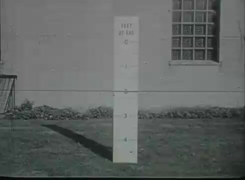 The still shows a marked pole in a grassy area, indicating measurements in feet, likely used for reference in the installation or maintenance of telephone wires. The background features a building with a window, and the overall scene is presented in a monochrome aesthetic typical of late 1940s to early 1950s films. The image is straightforward, focusing on the measurement criteria related to telephone drop wire installation.