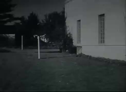 The still from 'Bell System Film - Drop Wire Runs Part 1 (b)' shows an exterior scene focused on a building, likely a Central Office. In the foreground, there are utility poles or posts indicating points for telephone wire connections. The environment appears to be grass and possibly shrubbery, suggesting a suburban setting. The film likely illustrates the steps involved in connecting telephone wires from the poles to residential or business customers. The overall tone reflects the technical and instructional nature of the film, typical of the late 1940s to early 1950s.