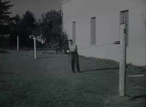 The still depicts a scene from the Bell System film 'Drop Wire Runs Part 1 (b),' illustrating the process of installing telephone wire. A man stands in an open area near a utility pole, possibly preparing to connect or adjust the wire. The background features a building and a few other poles, emphasizing a rural or suburban setting. The visual focuses on the practical steps involved in bringing telephone service from a central office to the customer.