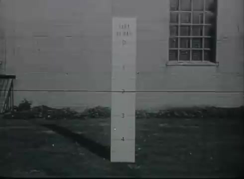 The still from the Bell System film shows a vertical sign or post marked with numerical indicators on a grassy area next to a building. The white post serves as a reference point with measurements (marked from 1 to 4) that may relate to telephone drop wire installation. In the background, there is a window and some equipment likely related to telephone service, emphasizing the technical context of connecting telephone lines from the central office to customers. The film focuses on instructional elements relevant to telephone line installation practices of the late 1940s to early 1950s.