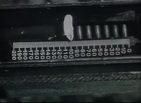 The still features a close-up of a measurement tool or gauge, likely used to read precise distances or measurements related to telephone wire installation. The scale is marked in units, with clear divisions indicating measurements from 0 to 200. This would be relevant in the context of the film, which focuses on the technical aspects of installing drop wires from telephone poles to customer premises. The precision shown in the measurement aligns with the film's educational intent to explain the process methodically.