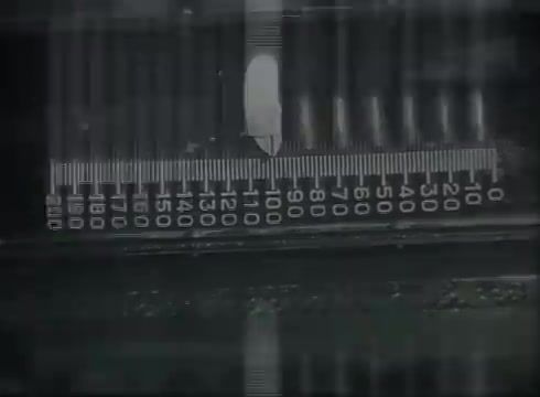 The still features a close-up of a measuring tool, likely a ruler or scale, with clearly marked increments. The image is presented in a monochrome style typical of 16mm films from the late 1940s or early 1950s. This specific segment likely illustrates a step in the process of measuring or laying telephone wire, emphasizing precision critical in connecting telephone lines from utility poles to customer premises within the context of Bell System operations.