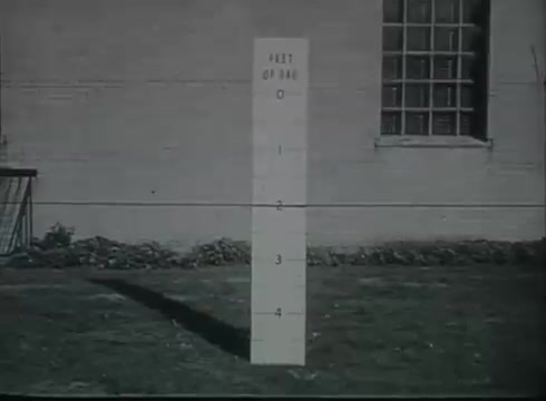 The still features a measuring post marked in feet, indicating heights from 0 to 4 feet, placed on a grassy area next to a building. This visual context is likely used to demonstrate the proper height for telephone drop wire installation, a key element in connecting the telephone line from utility poles to customers' locations. The film's educational aim is to provide clear guidelines for technicians working with telephone infrastructure.