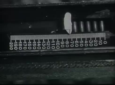 The still shows a close-up of a measuring device, likely used in the context of managing or installing telephone wires. It features a marked scale, indicating measurements, which may be related to wire lengths or installation parameters. The design suggests a technical focus typical of instructional materials from the late 1940s to early 1950s, aimed at demonstrating the steps involved in connecting telephone wires from utility poles to customer premises.