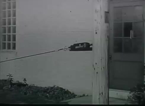 The still from the Bell System film depicts a telephone wire running from a utility pole to a building. It shows the wire extending toward a door, likely indicating the connection point for telephone service. The setting is utilitarian, reflecting the era of the late 1940s to early 1950s, focusing on the practical aspects of telecommunications infrastructure. The image captures a moment in the step-by-step process of connecting telephone service to customers.