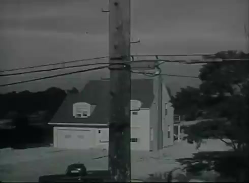 The still from 'Bell System Film - Drop Wire Runs Part 1 (b)' shows a telephone utility pole with several wires running down to a nearby house. The scene is set in a suburban area, with the house positioned prominently in the background. The pole is central to the composition, showcasing how telephone wiring connects from the pole to the customer’s premises. The film captures the infrastructure involved in telephone communication during the late 1940s to early 1950s, highlighting the practical aspects of wire installation. The overall tone is instructional, aimed at demonstrating telephone service delivery.