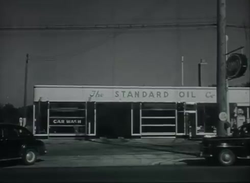The still captures a scene from a late 1940s to early 1950s Bell System film. It features a Standard Oil service station, identifiable by its signage. The black-and-white image shows a car wash area and several parked cars. Utility poles with overhead wires are visible, reflecting the context of telephone wire installation. The composition emphasizes the intersection of infrastructure and customer service during that era.