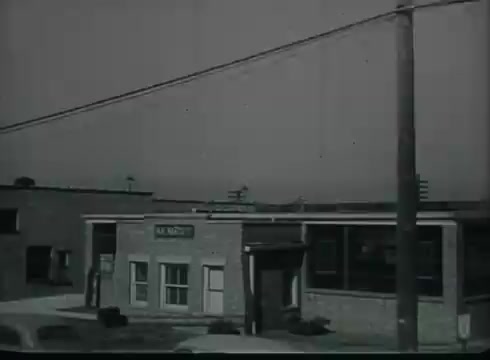The still from 'Bell System Film - Drop Wire Runs Part 1 (b)' depicts a brick building labeled 'A.L. Nelson,' likely a local business or central office. In the foreground, there’s a telephone pole with wires extending overhead, illustrating the connection from the pole to the structure. The scene captures a utilitarian view of telecommunications infrastructure from the late 1940s to early 1950s, focusing on the process of delivering telephone service to consumers.