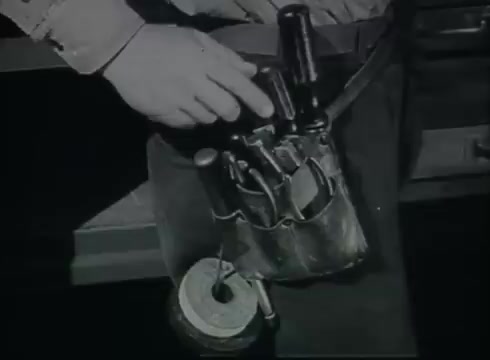 The still depicts a close-up of a technician's tool belt featuring various tools essential for telephone wire installation. A hand is shown interacting with the tools, highlighting their arrangement and functionality. The context relates to the processes and equipment involved in connecting telephone lines from utility poles to customers, as described in the Bell System film from the late 1940s to early 1950s.