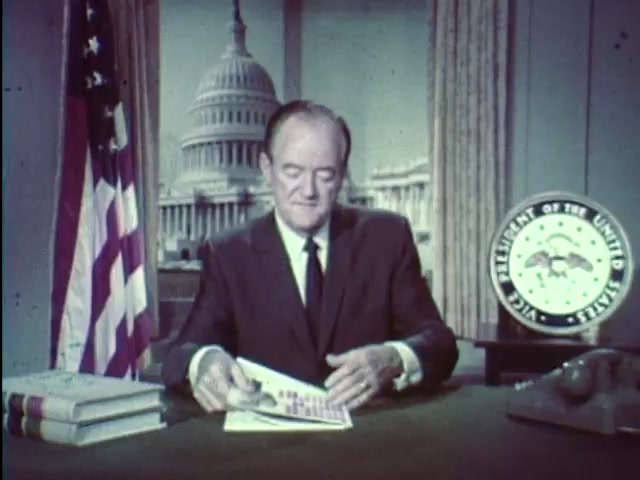 The still features Vice President Hubert Humphrey seated at a desk, addressing viewers in front of a backdrop that includes the U.S. Capitol building. He is dressed in a suit and appears to be writing or reviewing documents. To his left, there are two books stacked, and on his right is a vintage telephone and the presidential seal. The image conveys a sense of official government communication, emphasizing an educational message to encourage students to stay in school.