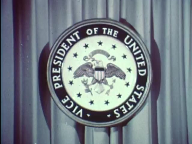 The still features the official seal of the Vice President of the United States, prominently displayed against a backdrop of draped fabric. The seal showcases an eagle holding arrows and an olive branch, emphasizing themes of strength and peace. This image serves as a setting for a public service announcement from 1967, in which Vice President Hubert Humphrey encourages students to stay in school, highlighting the importance of education. The overall tone conveys authority and commitment to educational values.