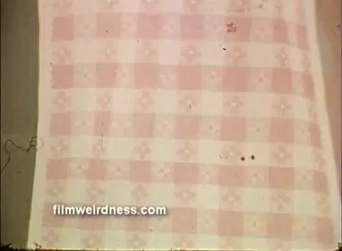 The still features a close-up of a patterned fabric, likely a napkin, depicting a pink checkered design with small floral motifs. There are some film artifacts, such as dust or scratches, indicating its vintage nature. This imagery relates to a commercial for Mardi Gras-themed napkins.