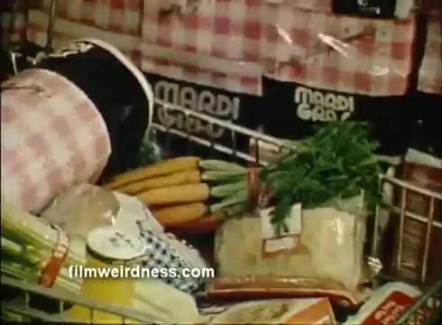 The still features a grocery cart filled with various colorful items, including fresh vegetables like green onions and carrots, alongside packaged goods. The background showcases stacks of Mardi Gras napkins, emphasizing the commercial's focus on this product. The overall scene conveys a festive shopping atmosphere typical of Mardi Gras celebrations.