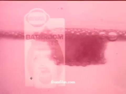 The still features a shot of a bubbling reaction in water, showcasing an Airwick Bathroom tablet in a pink-tinted environment. The focus is on the dissolving tablet, creating a dynamic visual, possibly emphasizing its effectiveness in cleaning or deodorizing. Bubbles rise around the product label, enhancing the promotional aspect of the ad. The overall aesthetic is reflective of 1970s advertising styles, combining vibrant colors with product demonstrations.