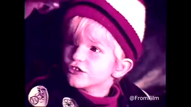 The still from the C&P Telephone commercial features a young child with blond hair, wearing a striped hat and a jacket adorned with patches. The background appears slightly blurred, emphasizing the child’s expressive face as they engage in conversation. The scene likely conveys a sense of warmth and connection, aligning with the tagline “The next best thing to being there.” The commercial reflects the era's charm and promotes the idea of communication through the Bell System.