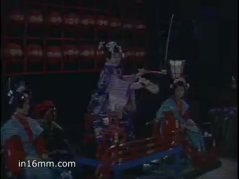 The still from 'Pan Am Far East Special - 1960s' likely features performers in traditional attire, possibly depicting a cultural performance or theatrical event. The composition suggests a focus on a central performer with others seated, indicating a setting typical of theatrical or entertainment presentations from that era. The vibrant colors and costumes reflect the stylistic choices of the time, capturing a moment that highlights cultural representation in travel promotional footage.   To help pinpoint a more specific date, consider exploring the costumes, stage design, and any identifiable features in the performance style, as these can provide clues to the time period.
