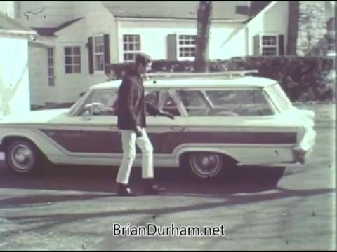 The still depicts a young man in a casual outfit walking towards a vintage station wagon parked on a driveway. The scene appears to be set in a suburban environment, with houses visible in the background. The overall tone is nostalgic, reflecting a 1960s aesthetic.
