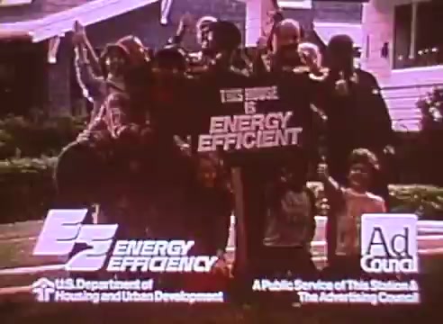 The still features a diverse group of adults and children joyfully posing in front of a house, prominently displaying a sign that states “This House is ENERGY EFFICIENT.” The image emphasizes community engagement in energy conservation, supported by logos from the U.S. Department of Housing and Urban Development and the Ad Council, highlighting it as a public service announcement.