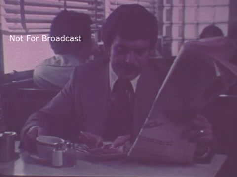 The still from the 1975 'Job Service Ohio' PSA features a man sitting in a diner, reading a newspaper while having a meal. The setting conveys a casual atmosphere, possibly suggesting the theme of job searching or employment opportunities. The color tone is muted, characteristic of the period's film style. The phrase 'Not For Broadcast' is overlayed, indicating that this image is not for public distribution.