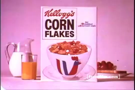 The still features a vintage Kellogg's Corn Flakes box prominently displayed against a pastel background. In front of it, there's a bowl of corn flakes topped with milk. A glass of orange juice and a pitcher are visible alongside a plate of grapes and a slice of toast, all reflecting a wholesome breakfast scene typical of the 1970s. The overall aesthetic is colorful and nostalgic.